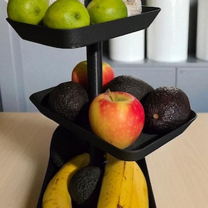 May include: A black, three-tiered serving stand filled with fresh fruit. Limes, apples, avocados, and bananas are arranged on the trays. The stand is on a light-colored wooden surface, with white pots and plants in the background.
