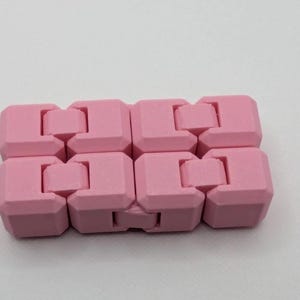 May include: A pink fidget toy composed of interlocking, cube-shaped pieces. The toy is designed to be manipulated and moved, offering a tactile experience. The color is a soft, pastel pink, and the toy is shown against a white background.