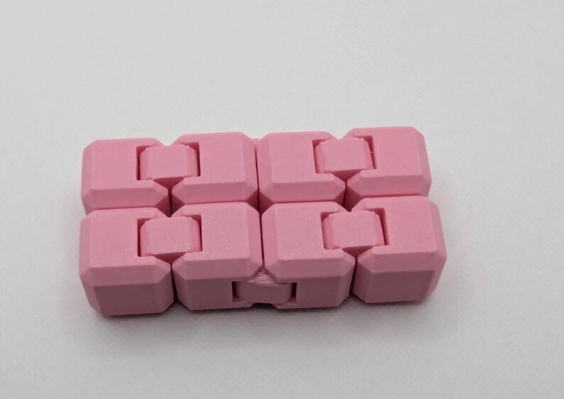 May include: A pink fidget toy composed of interlocking, cube-shaped pieces. The toy is designed to be manipulated and moved, offering a tactile experience. The color is a soft, pastel pink, and the toy is shown against a white background.
