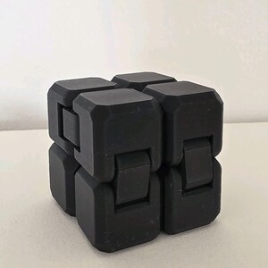 May include: A black fidget cube toy. The cube is made of several connected blocks that can be manipulated. The toy is designed to relieve stress and improve focus. The cube is sitting on a white surface.