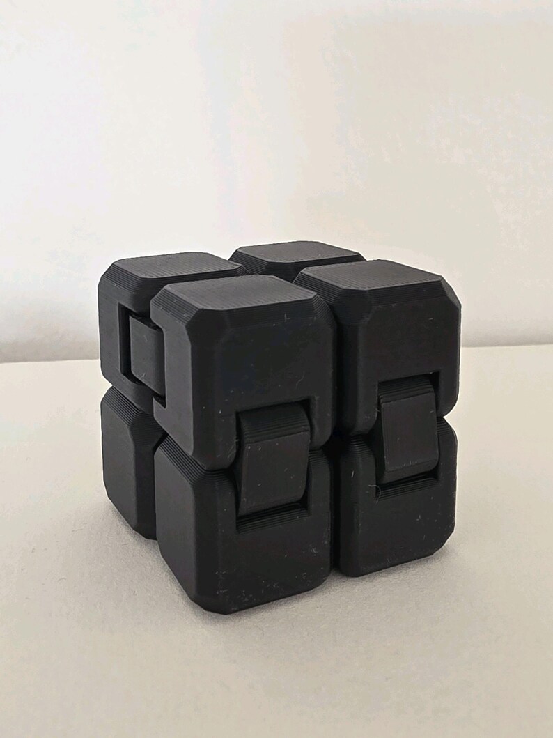 May include: A black fidget cube toy. The cube is made of several connected blocks that can be manipulated. The toy is designed to relieve stress and improve focus. The cube is sitting on a white surface.