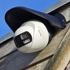 May include: White and black security camera mounted on weathered wooden siding, under a black protective hood. The camera has the brand name "annke" printed on its side. The background is a clear blue sky.