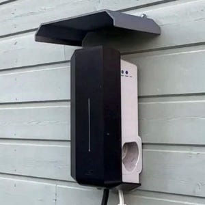 Puede incluir: Un cargador de vehículos eléctricos montado en una pared de madera verde claro. El cargador tiene una parte delantera rectangular negra y un lado blanco con un puerto de carga. Se instala una marquesina de metal gris oscuro encima del cargador.