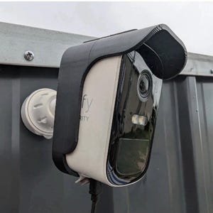May include: A white and black security camera with a black visor is mounted on a gray metal surface. The camera has the word "eufy" printed on it. A white mounting bracket is visible.