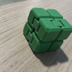 May include: A green fidget cube composed of interlocking blocks. The cube is made of a solid green material and sits on a light wood-grain surface. The cube's design allows for manipulation and movement of its individual parts.