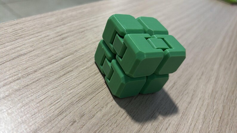 May include: A green fidget cube composed of interlocking blocks. The cube is made of a solid green material and sits on a light wood-grain surface. The cube's design allows for manipulation and movement of its individual parts.