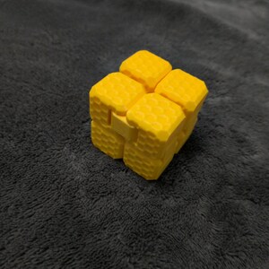 May include: A bright yellow fidget cube with a honeycomb-like texture. The cube is made of several connected blocks that can be manipulated. The background is a dark gray, textured surface.