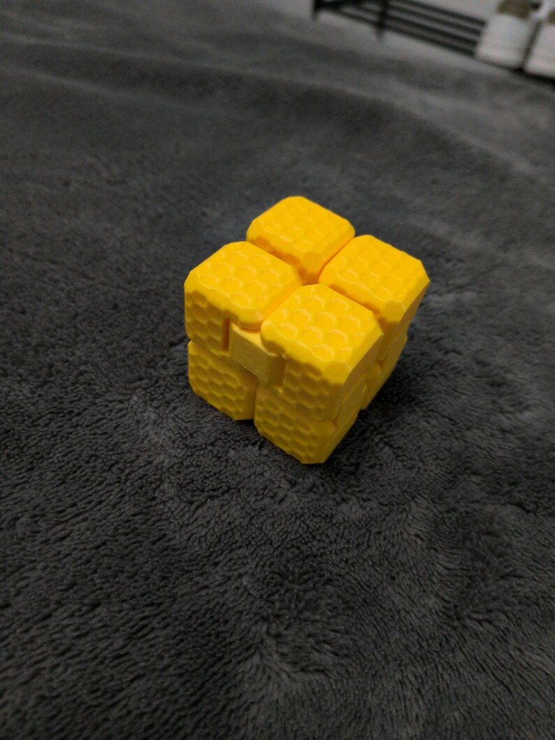 May include: A bright yellow fidget cube with a honeycomb-like texture. The cube is made of several connected blocks that can be manipulated. The background is a dark gray, textured surface.