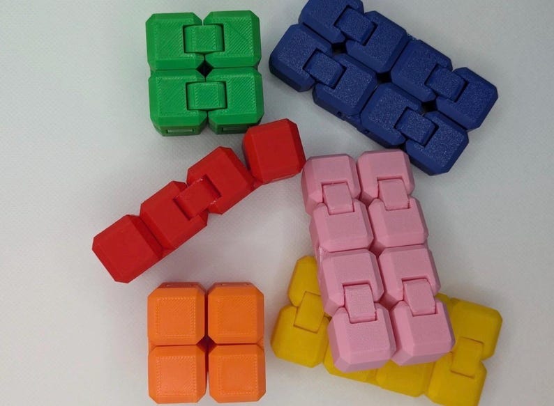 May include: An assortment of colorful fidget toys in green, blue, red, pink, orange, and yellow. The toys are made of connected blocks that can be manipulated. The image is taken from a high angle, with the toys arranged on a white surface.