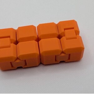 May include: An orange fidget toy, composed of interconnected, rectangular blocks. The blocks are a vibrant, solid orange color and are designed to move and flex. The toy is shown against a white background.