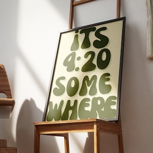 May include: A framed print with a light yellow background and green text that reads "ITS 4:20 SOMEWHERE". The print is sitting on a wooden easel.