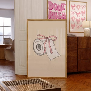 Könnte beinhalten: Eine gerahmte Illustration einer Toilettenpapierrolle mit einem rosa Band darum gebunden. Die Illustration ist auf einem beigefarbenen Hintergrund und der Rahmen ist aus hellem Holz. Der Text "Don't Rush" ist im Hintergrund sichtbar.