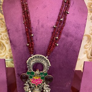 Op de afbeelding: Een ketting met meerdere strengen kleine, donkerrode kralen. De zilverkleurige hanger heeft een ingewikkeld filigraanontwerp, groene en rode details en een roze lotusbloem in het midden. De ketting wordt getoond op een paarse etalagepop.