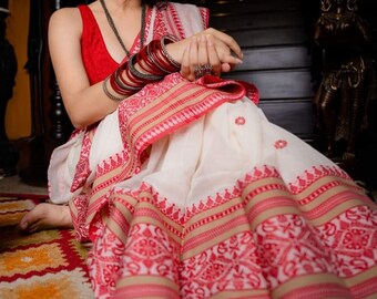 May include: A white saree with red and gold trim and intricate patterns. The saree is draped around a person and has a red sleeveless blouse.