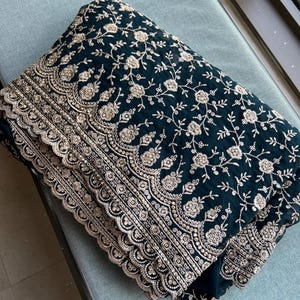 Peut inclure: Tissu bleu canard foncé avec broderie florale dorée complexe. Le tissu plié met en valeur le motif détaillé et les bords festonnés. Le motif présente des fleurs et des vignes délicates, créant un textile luxueux.