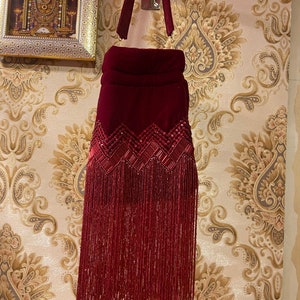 Puede incluir: Un bolso de mano de terciopelo granate con flecos de cuentas y una correa a juego. El bolso tiene un diseño acolchado y está decorado con lentejuelas.