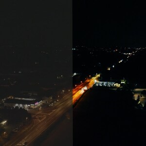 May include: A night time aerial view of a city with a highway running through the center. The highway is lit up with streetlights and car headlights. The city is mostly dark, but there are some lights from buildings and houses.