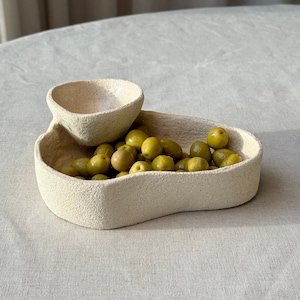 May include: A textured, off-white ceramic serving dish with two connected compartments. The larger section is filled with green olives. The smaller, elevated section is empty. The dish has a unique, organic shape.
