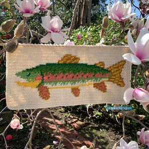 May include: A crocheted wall hanging featuring a colorful fish design. The fish is green, pink, and yellow with a white background. The hanging is displayed against a backdrop of pink magnolia blossoms.