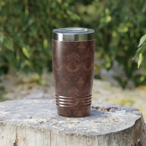 May include: A brown and silver stainless steel tumbler with a wood grain pattern sits on a tree stump.