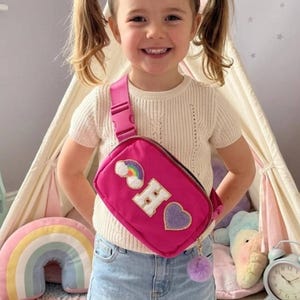 Puede incluir: Una niña sonríe, usando una riñonera rosa con un parche de arcoíris y corazón. La bolsa tiene un pompón morado y la letra "H". Lleva una camisa blanca y una falda vaquera. Una tienda tipi y un oso de peluche están en el fondo.