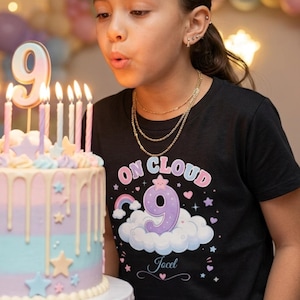 May include: A young person blowing out candles on a birthday cake. The cake is decorated with pastel colors, stars, and a large number 9 candle. The person is wearing a black t-shirt with the text "ON CLOUD 9" and a cloud graphic.