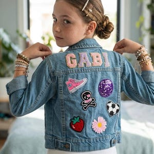 May include: A light blue denim jacket with patches, including a pink high-top sneaker, a skull and crossbones, a soccer ball heart, and a strawberry. The name "GABI" is embroidered on the back in pink letters.