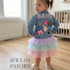 May include: A young girl wearing a denim jacket with patches and a multi-colored tutu skirt. The jacket has the name "KATIE" in pink letters on the back. Patches include a star, a heart, a unicorn, and a bow. The text "SEWN-ON PATCHES" is at the bottom.