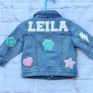 Denim jacket for a little girl with the name Leila on the back in white chenille letters with gold glitter around the letters. There are other patches of a paw heart, star, lightning and flowers in pastel colors.