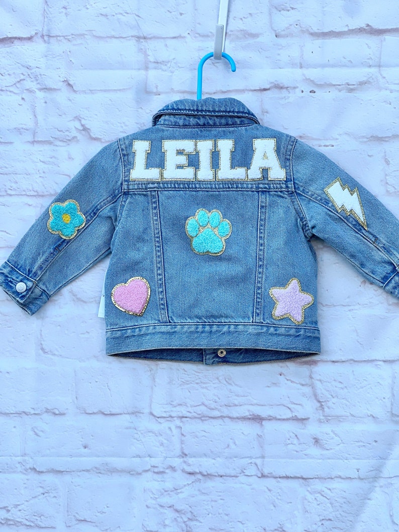 Denim jacket for a little girl with the name Leila on the back in white chenille letters with gold glitter around the letters. There are other patches of a paw heart, star, lightning and flowers in pastel colors.