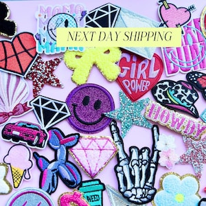 May include: A collection of colorful embroidered patches in various shapes, including stars, hearts, diamonds, and a smiley face. The patches feature glitter, text, and a skeleton hand. The background is a light pink color.