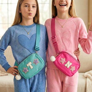 May include: Two young people wearing matching blue and pink outfits, each with a crossbody bag. The blue bag has a star and letter design, while the pink bag has a star and letter design. Both bags have a white pom-pom.