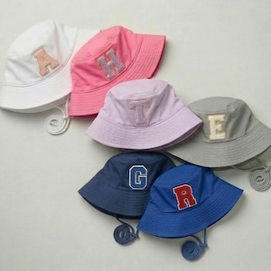 Può includere: Una collezione di cappelli a secchiello in vari colori, tra cui bianco, rosa, lavanda, grigio, blu navy e blu. Ogni cappello presenta una lettera ricamata diversa: A, H, T, E, G e R. I cappelli hanno un cordino per regolare la vestibilità.