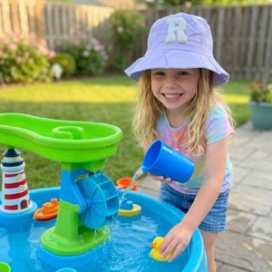 May include: A young girl smiles while playing with a colorful water table in a backyard. The water table features a lighthouse, a water wheel, and small toy boats. She is wearing a tie-dye shirt and a lavender bucket hat.