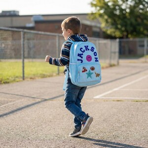 Puede incluir: Una mochila azul claro con el nombre "JACKSON" en letras azules y una carita sonriente. La mochila tiene parches de una estrella, un balón de fútbol y otros diseños. La persona lleva una camisa a rayas y vaqueros azules.