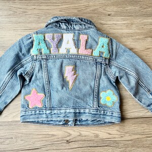 A medium/light wash denim jacket with the name AYALA written in chenille patches in pastel colors. There is also a pink star patch, a purple lightning patch and a blue flower patch. All patches have gold glitter outline.