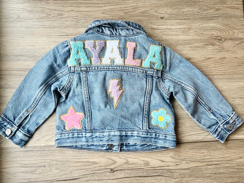 A medium/light wash denim jacket with the name AYALA written in chenille patches in pastel colors. There is also a pink star patch, a purple lightning patch and a blue flower patch. All patches have gold glitter outline.