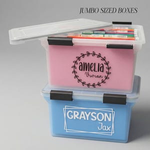 May include: Two clear plastic storage boxes, one pink and one blue, stacked on top of each other. The pink box has the name "Amelia" in a wreath design. The blue box has the name "Grayson Jax". The text "JUMBO SIZED BOXES" is above the boxes.