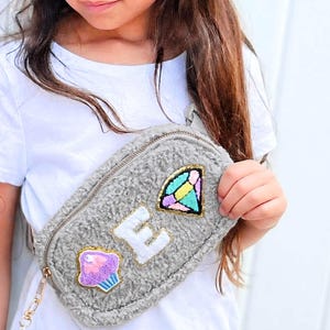 May include: A gray, fuzzy fanny pack with a gold zipper and chain. The pack features colorful patches, including a cupcake, a diamond, and the letter "E". The bag is worn across the body.