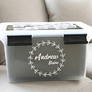 May include: A clear plastic storage bin with a white lid and black latches. The bin has a gray front with a white floral wreath design and the name "Andrew Blaine". Labeled file dividers are visible inside.