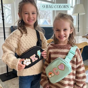 以下が含まれることがあります： 装飾パッチが付いた黒とミントグリーンのファニーパックを着用した2人の若い女の子。黒いバッグには足跡、「K」の文字、稲妻があります。ミントグリーンのバッグには「ACO」の文字、ハート、虹があります。「9 BAG COLORS」というテキストが上部にあります。