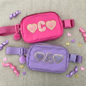 May include: Two fanny packs, one pink and one purple, with gold glitter letters and heart appliques. The pink pack has the letters "C O" and the purple pack has the letters "A D". Both have matching straps and pom-poms.