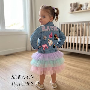 May include: A child in a denim jacket adorned with patches and a layered pastel tutu skirt. The jacket features the name "KATIE" and decorative patches. The image includes the text "SEWN-ON PATCHES" at the bottom.
