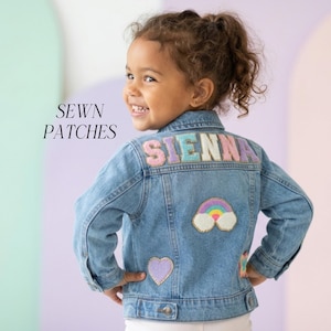 May include: A light blue denim jacket with the name "SIENNA" spelled out in colorful, sewn-on patches across the back. The jacket also features a rainbow, heart, and other decorative patches. The words "SEWN PATCHES" are printed on the left side.