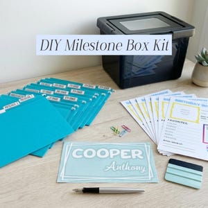 Puede incluir: Un kit de caja de hitos DIY con un contenedor de almacenamiento negro, carpetas de archivos turquesas y hojas de entrevista de cumpleaños. El kit incluye una tarjeta con el nombre, un bolígrafo y una pequeña planta suculenta en una maceta blanca. El texto "DIY Milestone Box Kit" es visible.
