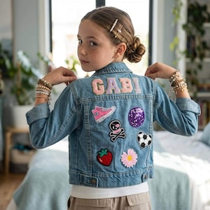 May include: A light blue denim jacket with patches, including a pink shoe, skull and crossbones, and a soccer ball heart. The name "GABI" is embroidered on the back in pink letters. The jacket is worn with bracelets.