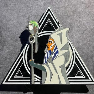 May include: Enamel pin featuring a character with orange skin, blue and white markings, and a white hooded cloak. The character holds a staff with a bird on top, set against a black and white triangle.