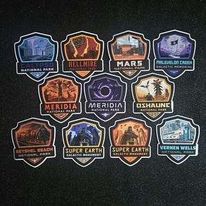 May include: A collection of shield-shaped stickers featuring various landscapes and text. The stickers include the names "Calypso National Park", "Hellmire National Park", "Mars National Park", and "Malevelon Creek Galactic Memorial". Other stickers say "Meridia National Park", "Oshaune National Park", "SeyShel Beach National Park", "Super Earth Galactic Monument", and "Vernen Wells National Park".
