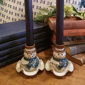 May include: Two snowman candle holders with black candles. The candle holders are made of ceramic and have a white glaze with blue and brown accents. The candles are lit and the flames are visible.