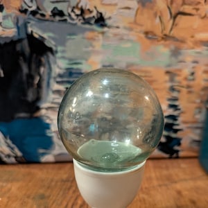 May include: A clear glass sphere sits atop a white ceramic egg cup. The sphere has a slight green tint and rests on a small, light green base. The egg cup has a rounded base and a slender stem.